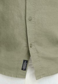 Chemise boutonnée vert olive en tissu texturé, avec deux boutons et une étiquette à l'ourlet. Conçue avec une coupe droite.