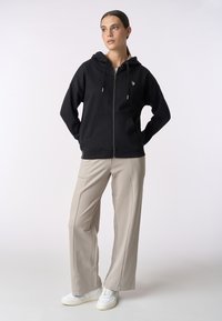 Felpa nera con zip, cappuccio, tasche frontali e hardware color oro, abbinata a pantaloni beige chiaro e sneakers bianche.