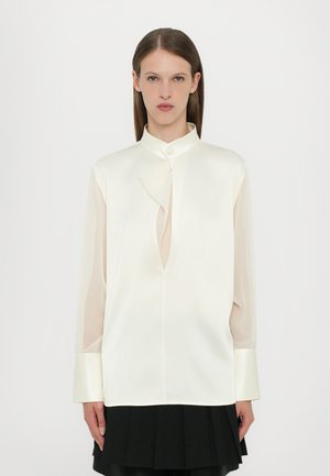 Chemise en soie off-white avec col mandarin, présentant un design de devant torsadé, des manches longues en tissu transparent et des poignets en satin, assortie d'une jupe plissée noire.