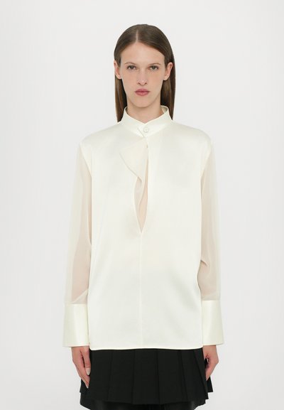 Chemise en soie off-white avec col mandarin, présentant un design de devant torsadé, des manches longues en tissu transparent et des poignets en satin, assortie d'une jupe plissée noire.