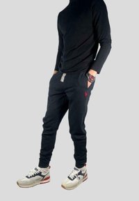 Černé tričko s dlouhými rukávy spojené s tmavě modrými tepláky s červeným logem, šedým šňůrkováním a bílými teniskami s modrými a červenými akcenty.