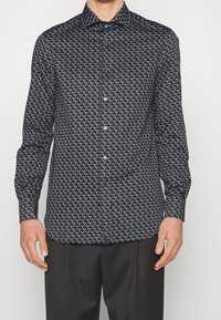 Černá košile s dlouhým rukávem, která má geometrický bílý vzor, zapínání na knoflíky a klasický límeček. Vyrobena z lehkého materiálu.