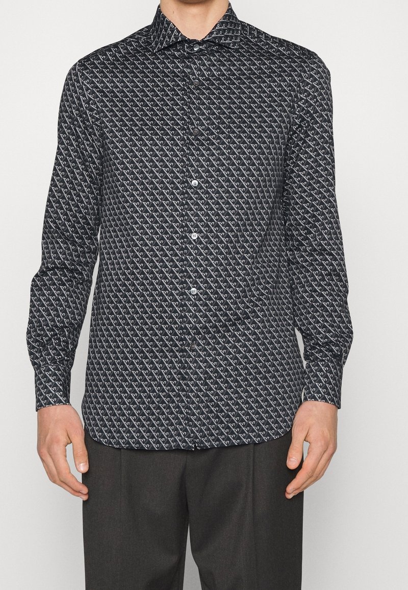 Černá košile s dlouhým rukávem, která má geometrický bílý vzor, zapínání na knoflíky a klasický límeček. Vyrobena z lehkého materiálu.
