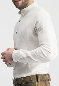 Weißes Leinenhemd mit Stehkragen, geknöpfter Leiste und langen Ärmeln, kombiniert mit strukturierten braunen Hosen.