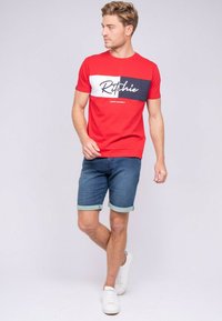 T-shirt en coton rouge avec une rayure horizontale bleu marine et blanche portant le logo "Ritchie", associé à un short en denim bleu avec des ourlets retournés. Baskets blanches.