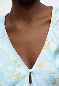 Ύφασμα πουκάμισου ανοιχτού γαλάζιου με κίτρινα και μπλε σχέδια. Διαθέτει V-λαιμόκοψη και ένα μικρό κουμπί πάνω από μια μικρή οπή.