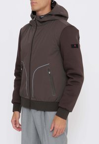 Uomo che indossa una giacca marrone con cappuccio e tasche con cerniera, e pantaloni grigi, in piedi davanti a uno sfondo bianco semplice.