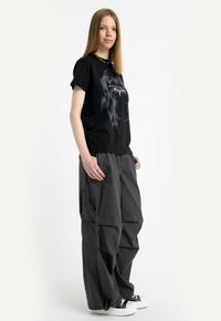 Młoda kobieta z prostymi włosami, ubrana w czarną koszulkę z grafiką, luźne ciemne spodnie cargo i czarno-białe sneakersy, stoi bokiem.