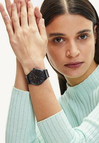 Zwarte analoge klok met een gaasband, ronde wijzerplaat en slanke uurmarkeringen. Draagt op een pols met lichte huid, gecombineerd met een mintgroene ribbeltrui.