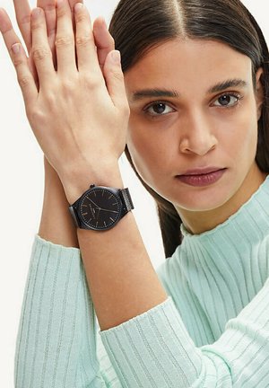 Schwarze analoge Uhr mit einem Netzband, rundem Zifferblatt und schlanken Stundenmarkierungen. Getragen an einem Handgelenk mit heller Haut, kombiniert mit einem mintgrünen, gerippten Pullover.