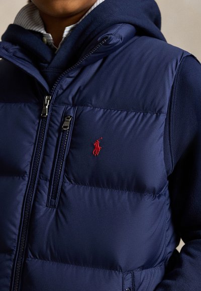 Primo piano di una persona che indossa un gilet trapuntato blu navy con un logo ricamato rosso di un giocatore di polo sopra una felpa con cappuccio blu navy.