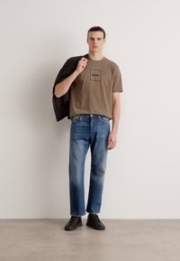 Brun bomull t-skjorte med svart logo, blå denimjeans og svarte sko. Grå blazer hengende over venstre arm; står foran en nøytral bakgrunn.
