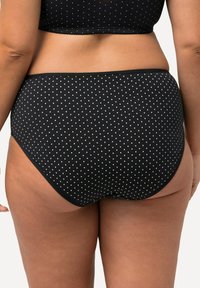 Zwarte hoogwaist slip met witte stippen, gemaakt van zacht materiaal en voorzien van een soepele elastische tailleband. Het achteraanzicht bied volledige bedekking.