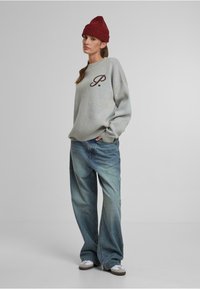 Sudadera gris de corte oversize con un logo marrón de "P.", combinada con unos jeans de mezclilla azul de corte holgado y un gorro de punto rojo. Zapatillas blancas y marrones.