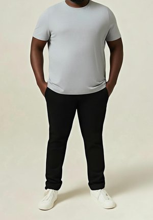 Mann trägt hellgraues kurzärmeliges T-Shirt, schwarze Hose und weiße Sneakers, steht mit Händen in den Taschen vor einem schlichten Hintergrund.