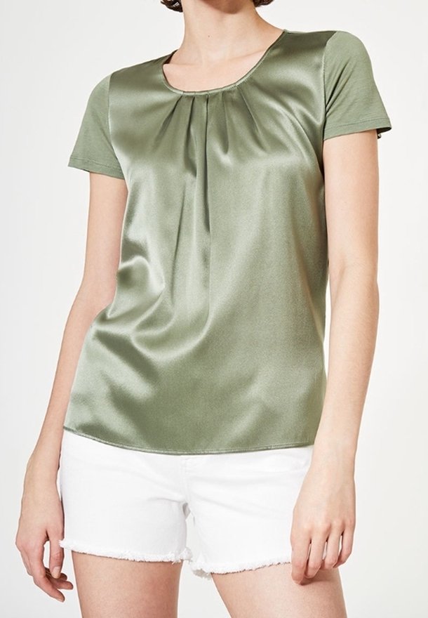 Femme portant un chemisier en satin vert olive à manches courtes avec des plis au niveau de l'encolure et un short en jean blanc effiloché.