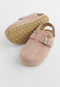 Sabots en daim rose avec une bride réglable, dotés d'une boucle métallique et d'une semelle en caoutchouc beige texturé avec un motif ondulé.