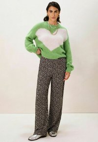 Pull en tricot vert avec un motif de cœur blanc, associé à un pantalon large à imprimé léopard et des chaussures métalliques argentées, portant des colliers superposés.