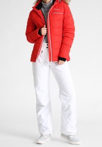 Chaqueta acolchada roja con capucha forrada de piel, cremallera frontal y puños negros; combinada con pantalones blancos de nieve y calzado gris claro.
