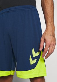 Hombre con pantalones cortos deportivos azul marino y amarillo neón con una camiseta negra, mostrando la mano derecha y parte del torso inferior y las piernas.