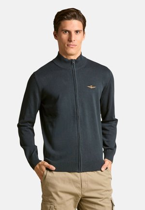 Maglione blu navy con cerniera e collo alto, realizzato in tessuto lavorato a maglia, con polsini e orlo a coste, e un piccolo logo ricamato sul petto.