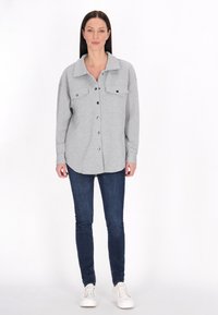 Camicia grigia con colletto, due tasche frontali e vestibilità rilassata, abbinata a jeans blu scuro e sneakers bianche.