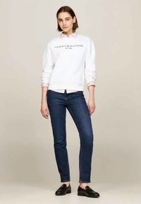 Λευκό βαμβακερό φούτερ με λογότυπο "TOMMY HILFIGER", συνδυασμένο με σκούρα μπλε στενά τζιν. Μοντέλο που φορά μαύρες loafers, που στέκεται σε ουδέτερη φόντο.