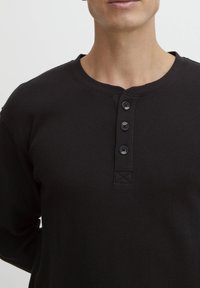 Črna teksturirana dolgocelana henley majica z okroglim izrezom, štirimi gumbi in všitimi X detajli pod gumbi.