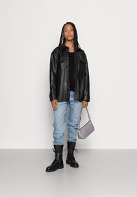Fekete bőr overshirt elülső zsebekkel, fekete felsőn viselve; világoskék bő szárú farmernadrág; fekete fűzős harci bakancs; szürke textúrált kézitáska.