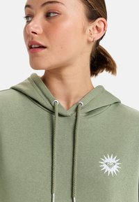Sudadera verde olivo con un cordón trenzado, que presenta un emblema gráfico de corazón blanco en un diseño de esplendor en el pecho.