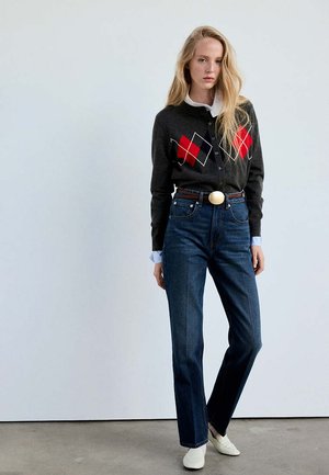Donna con capelli biondi lunghi indossa un cardigan grigio scuro con motivi a diamante rossi, jeans blu, camicia bianca e scarpe bianche, in piedi all'interno.