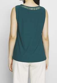 Top senza maniche color teal con un collo accentuato in pizzo, caratterizzato da una vestibilità comoda e una texture liscia, indossato con pantaloni di colore chiaro.