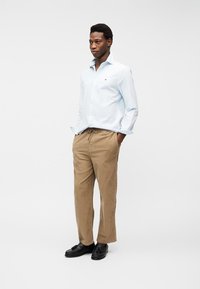 Νεαρός άνδρας στέκεται με τα χέρια στις τσέπες, φορά ανοιχτό μπλε πουκάμισο, μπεζ παντελόνι και μαύρα μοκασίνια, μπροστά σε απλό λευκό φόντο.