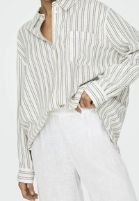Camicetta a righe bianca e marrone chiaro. Presenta una tasca, una vestibilità oversize e i polsini arrotolati. Abbinata a pantaloni bianchi larghi.