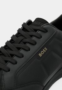 Black sneaker featuring textured synthetic material, thin laces, and a gold "BOSS" logo. The sole is ribbed for traction.