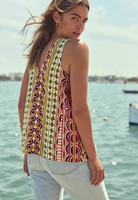 Haut sans manches avec un décolleté en V, présentant un motif géométrique audacieux en rouge, vert et jaune sur un fond blanc. Porté avec un jean bleu clair.