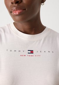 Kreminė marškinėlių su "TOMMY JEANS" ir "NEW YORK CITY" tekstu, su logotipu raudonos, baltos ir mėlynos spalvų. Apvalus iškirpimas ir medvilninis audinys.