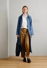 Vrouw in een lange blauwe denimjas, witte blouse, bruine broek, zwarte schoenen, met een grote zwarte tote bag, staand op een houten vloer.