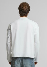 Weißes Langarm-T-Shirt mit Rundhalsausschnitt, glatter Textur und sichtbaren Nähten; kombiniert mit hellblauen Jeans mit Gesäßtaschen.
