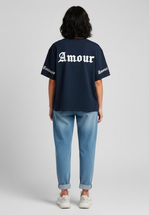 Navyblauwe oversized t-shirt met "Amour" in witte gotische letters op de achterkant en mouwen. Gecombineerd met lichtblauwe opgerolde jeans en witte sneakers.