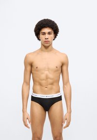 Μαύρα βαμβακερά εσώρουχα με λευκή ζώνη μέσης που φέρει το λογότυπο "TOMMY HILFIGER". Το μοντέλο στέκεται σε όρθια στάση μπροστά σε ουδέτερο φόντο.