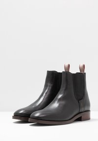 Bottines en cuir noir à finition lisse, avec des panneaux latéraux élastiques et des languettes de tirage contrastées. Talon en bois marron pour plus de stabilité.