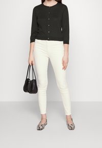 Dunkelgrüne, kurz geschnittene Strickjacke mit Knopfleiste, kombiniert mit weißen Slim-Fit-Jeans und einer schwarzen Handtasche. Die Schuhe haben ein Schlangenhautmuster.