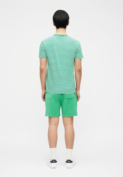 Άτομο που στέκεται πλάι και φοράει πράσινη και λευκή ριγωτή μπλούζα, πράσινα σορτς, λευκές κάλτσες και λευκά αθλητικά παπούτσια με φόντο λευκό.