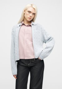 Νέα γυναίκα με μακριά ξανθά μαλλιά, που φοράει ένα γαλάζιο πλεκτό ζακέτα με κοτσίδες, ροζ πουκάμισο με υφή και σκούρο τζιν, στέκεται μπροστά σε απλό φόντο.