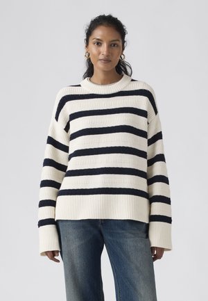 Mujer con pendientes de aro dorados, un suéter de punto a rayas blancas y negras, y jeans azules, de pie contra un fondo claro y liso.