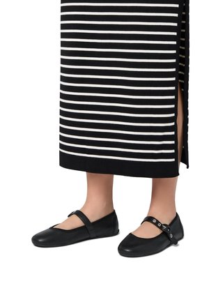 Zapatos planos negros con textura perforada, que cuentan con una correa con botones plateados. Se llevan con una falda midi a rayas en blanco y negro.