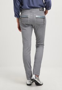 Šedé slim-fit džíny s hladkou texturou, s dekorativními zipy na zadních kapsách, v kombinaci s lesklými stříbrnými botami a modrým topem.