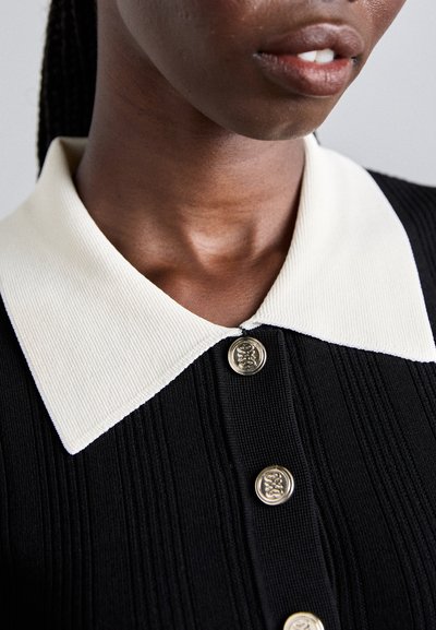 Primer plano de una persona que lleva una camiseta negra de canalé con un collar blanco en punta y botones decorativos dorados.