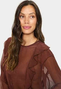 Brown, sheer blouse with ruffled detailing along the neckline and sleeves. Lightweight fabric with a smooth texture.
